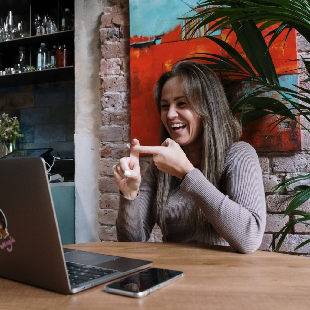 A woman laughs as she signs to someone on her laptop's video call, at a wooden table in front of a brick wall and a big palm. Best meeting of her week, and nobody had to say "you're on mute."