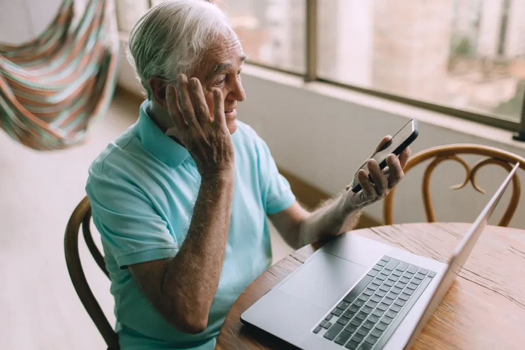 An older man in a turquoise polo presses a hand to his cheek and squints at the phone in his other hand, his laptop open on the table in front of him. He has just met a pop-up that won't close. You've been there too.